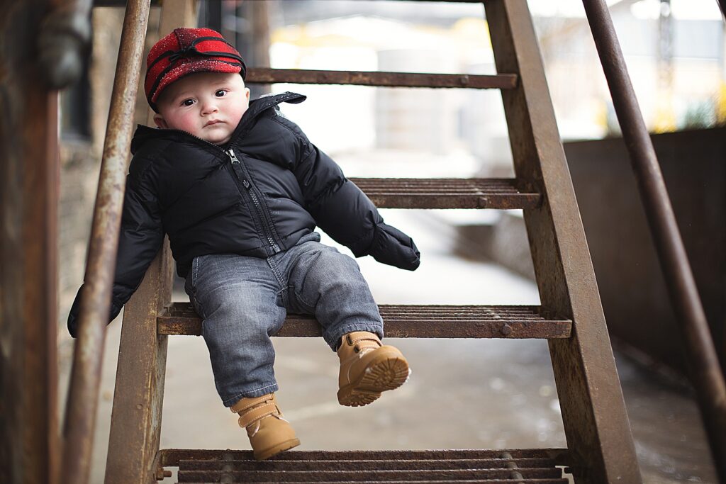 Little boy sitting on steps with a red buffalo hat and puffy jacket Minneapolis, MN
