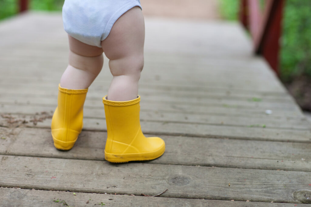 Close-up of a baby in bright yellow rain boots on a wooden path—a simple, seasonal detail. 