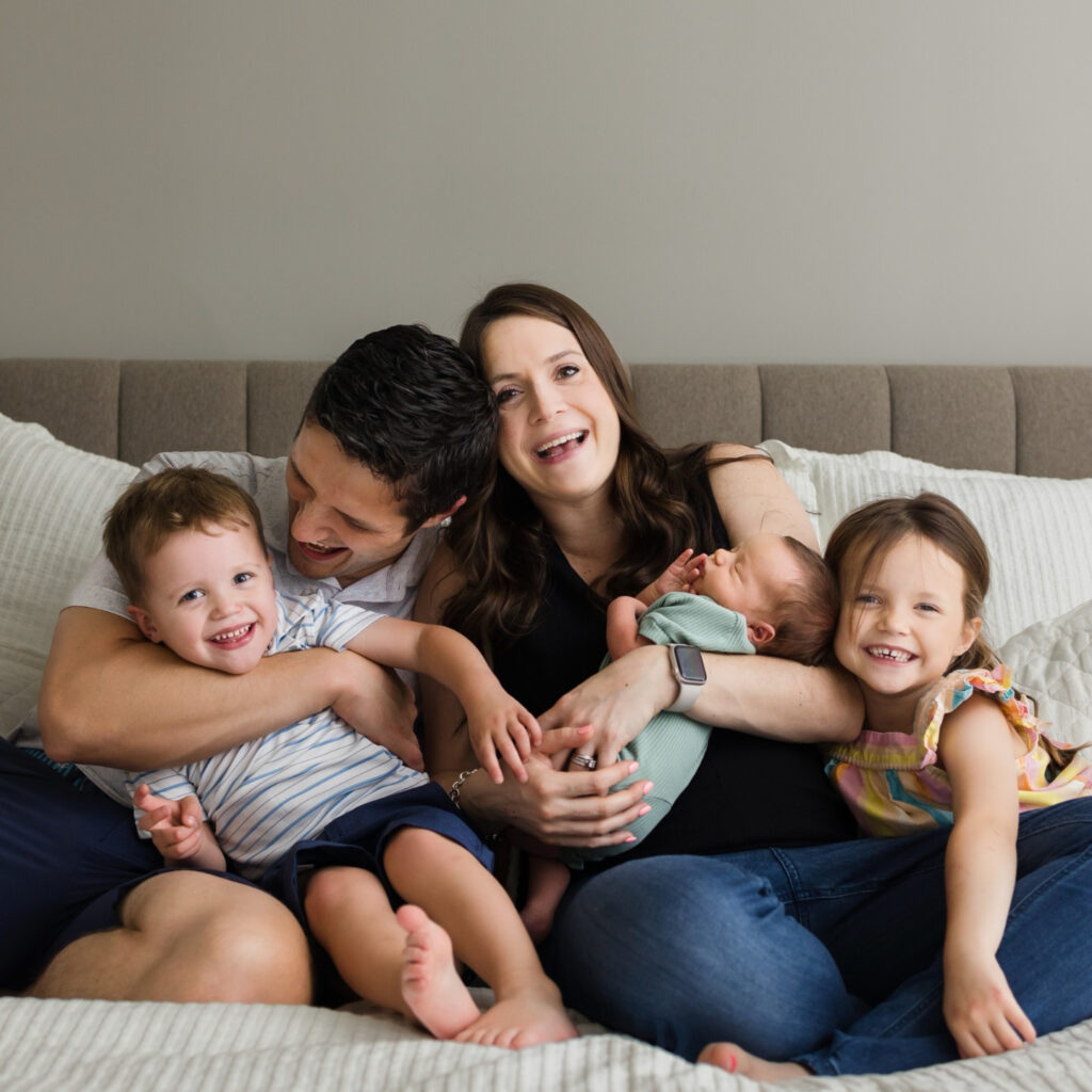 Family of five sitting together on bed, parents laughing while holding newborn, siblings smiling and cuddling close.