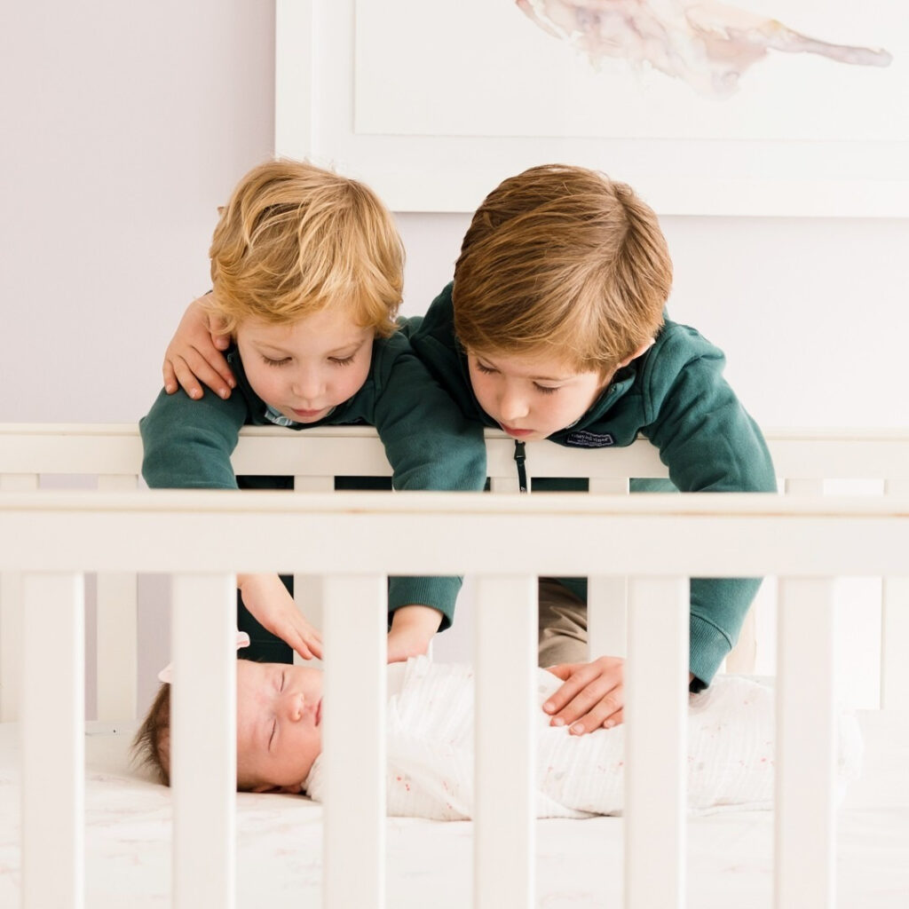 Two older brothers gently looking at newborn sibling in crib during a newborn and family photography session.
