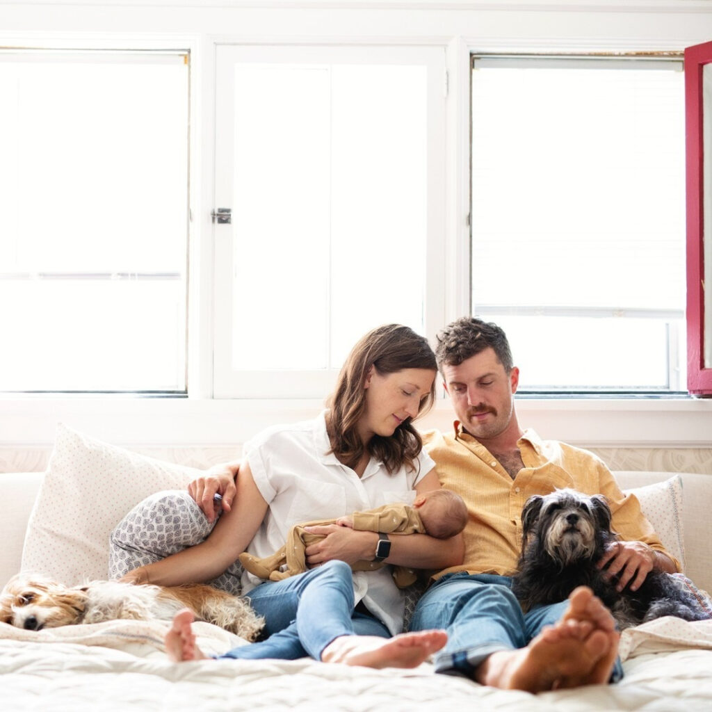 Parents snuggling newborn with two dogs on bed during a cozy newborn and family photography session.