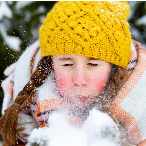 Young girl bundled in winter clothes blowing snow during winter photos, wearing a yellow knit hat and scarf.