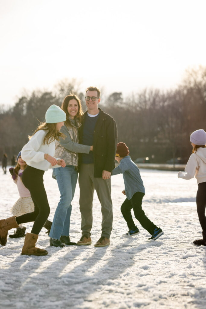 Parents smiling while children run and play around them on a snowy winter day.