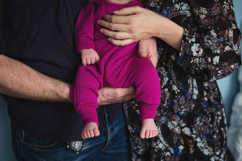 Baby in bright magenta romper held by parents, showing only hands, arms, and tiny feet.