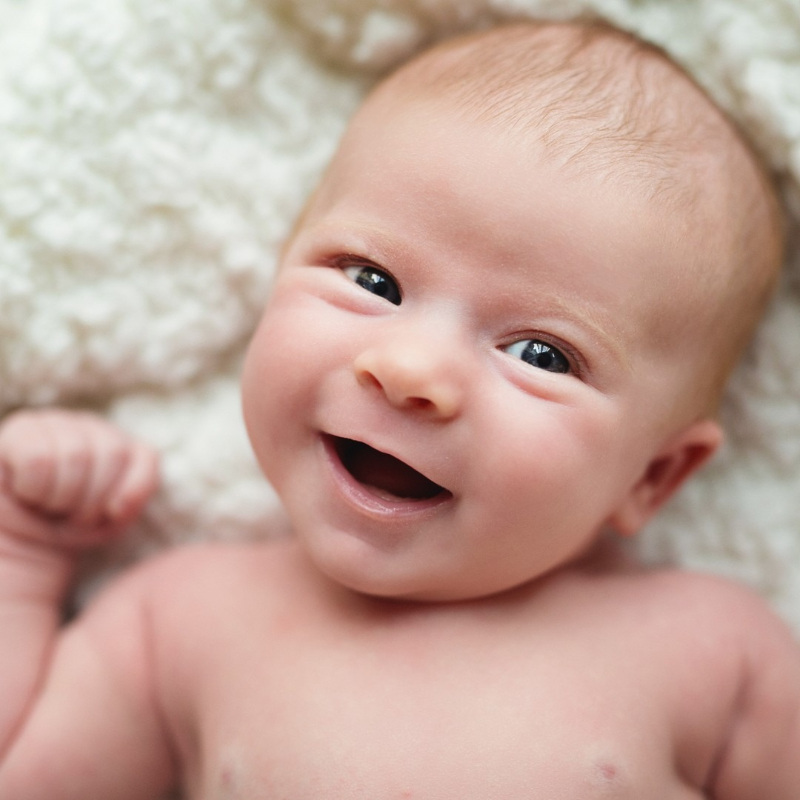 Smiling newborn lying on soft white blanket, eyes bright and fist raised in a playful pose.