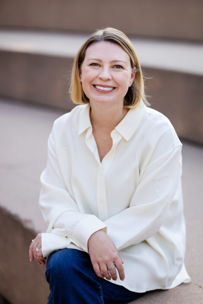 Woman in white blouse and jeans smiling during natural light headshot photography session outdoors.