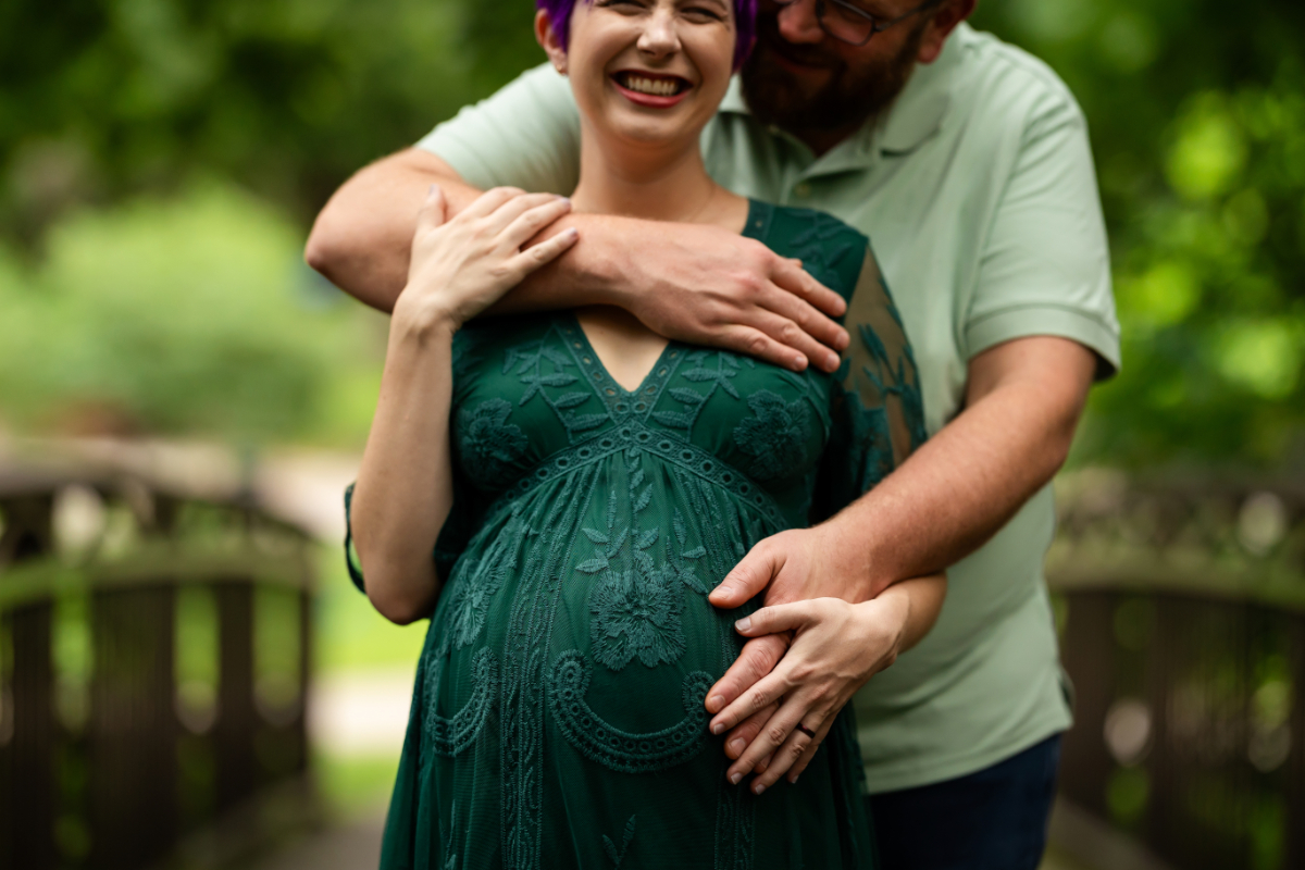 Pregnant couple standing on a wooded path, laughing as the partner wraps arms around the expectant mom, hands resting on her baby bump during a maternity session. | prenatal chiropractor