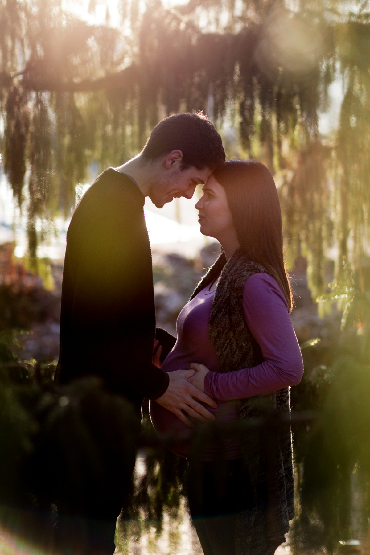 Expectant couple standing forehead to forehead near water at sunset, hands resting on a pregnant belly, sharing a quiet, emotional maternity moment.