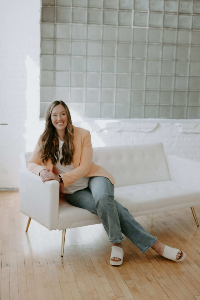Portrait of Hannah Lewis, a functional medicine doctor at Radiant Health Collective, smiling while seated in a bright, natural light setting. She wears a peach blazer and white top, representing the approachable, patient-focused care she offers women seeking root-cause health support.