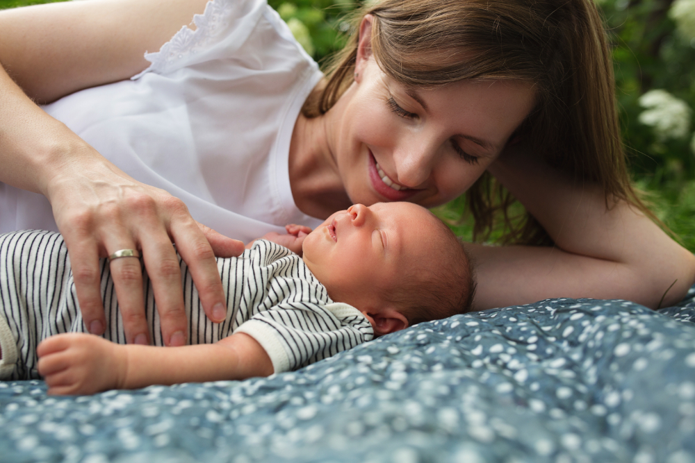Mother lying on a blanket outdoors, gently touching her sleeping newborn in a striped onesie, sharing a peaceful, sunlit moment together. | naturopathic doctor