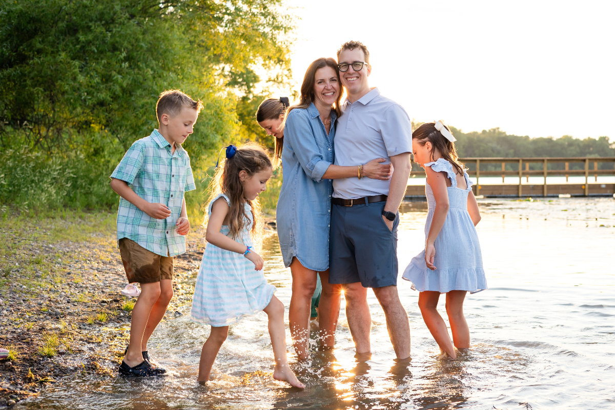 A family of six wading in a calm Minneapolis lake at golden hour, parents holding each other close while kids splash freely around them — exactly the kind of summer moment worth capturing.