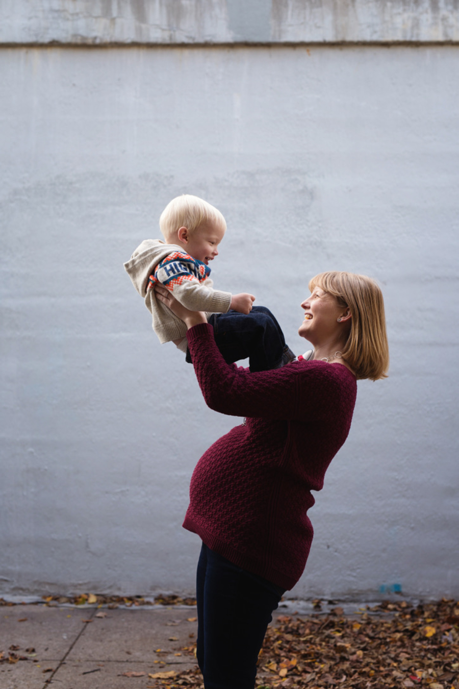 A pregnant mom in a burgundy sweater laughs as she lifts her smiling toddler into the air against a white brick wall during a fall maternity session.