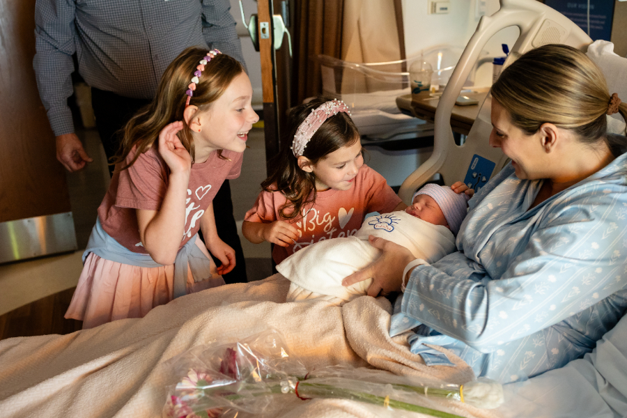 Two big sisters in matching pink tops lean in to meet their newborn sibling in mom's arms during a Fresh 48 hospital session in Minneapolis.