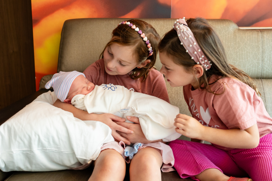 Two sisters with headbands sitting together, both gazing at the swaddled newborn baby between them during a Fresh 48 session with Megan Norman Photography | Advice for moms