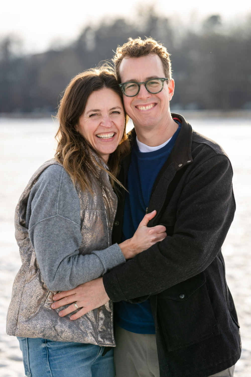 Happy couple embracing beside a calm lakeside during an outdoor portrait session in soft natural light. Both smile warmly at the camera while bundled in casual layers, creating a relaxed and intimate portrait with a peaceful winter backdrop.