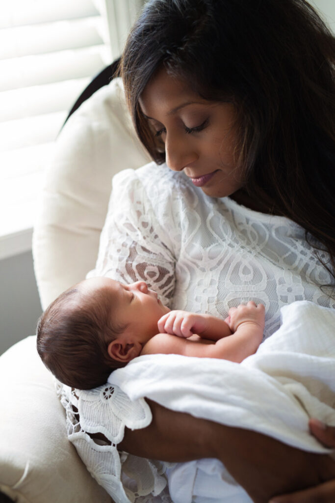 A mother gazing down at her sleeping newborn wrapped in white, the tender stillness that night nurse support helps protect in those early postpartum days.