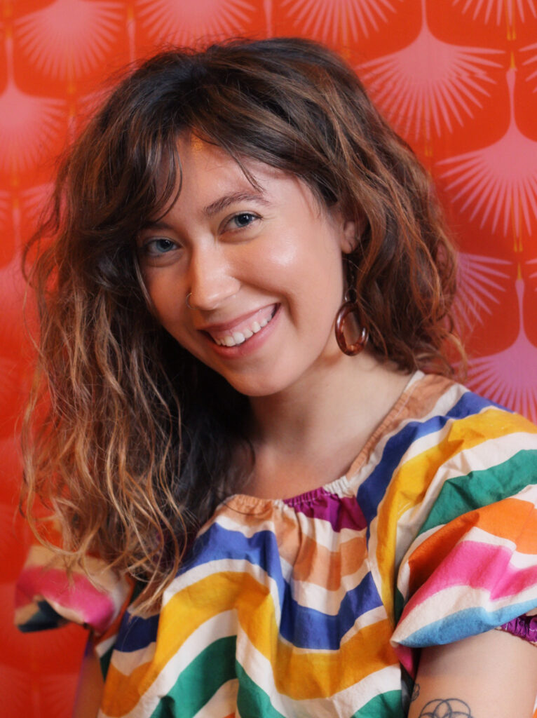 Hannah Kuduk, Minneapolis-based night nurse and postpartum doula, founder of The Folk Doula, smiling in a colorful top against a vibrant orange backdrop.