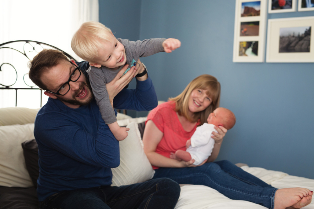 Dad holding toddler son up playfully while mom holds newborn baby on bed, capturing the joyful chaos of the first week home with a newborn, Minneapolis family photography.