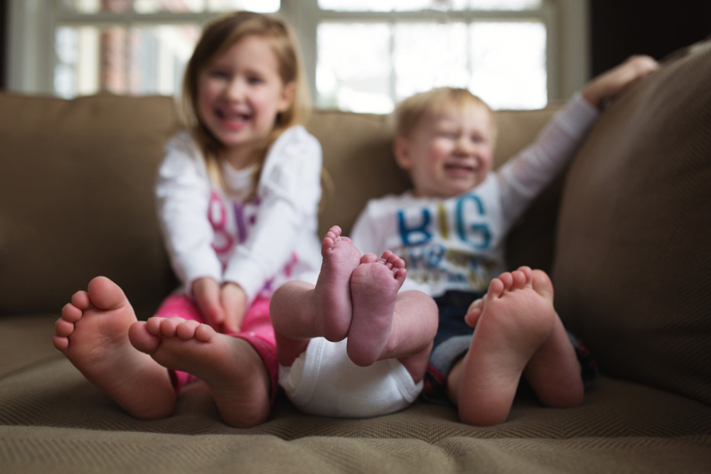 Two laughing siblings show off bare feet next to their newborn sibling's tiny feet on the couch, a sweet sibling moment during the first week home with a newborn, Minneapolis family photographer.