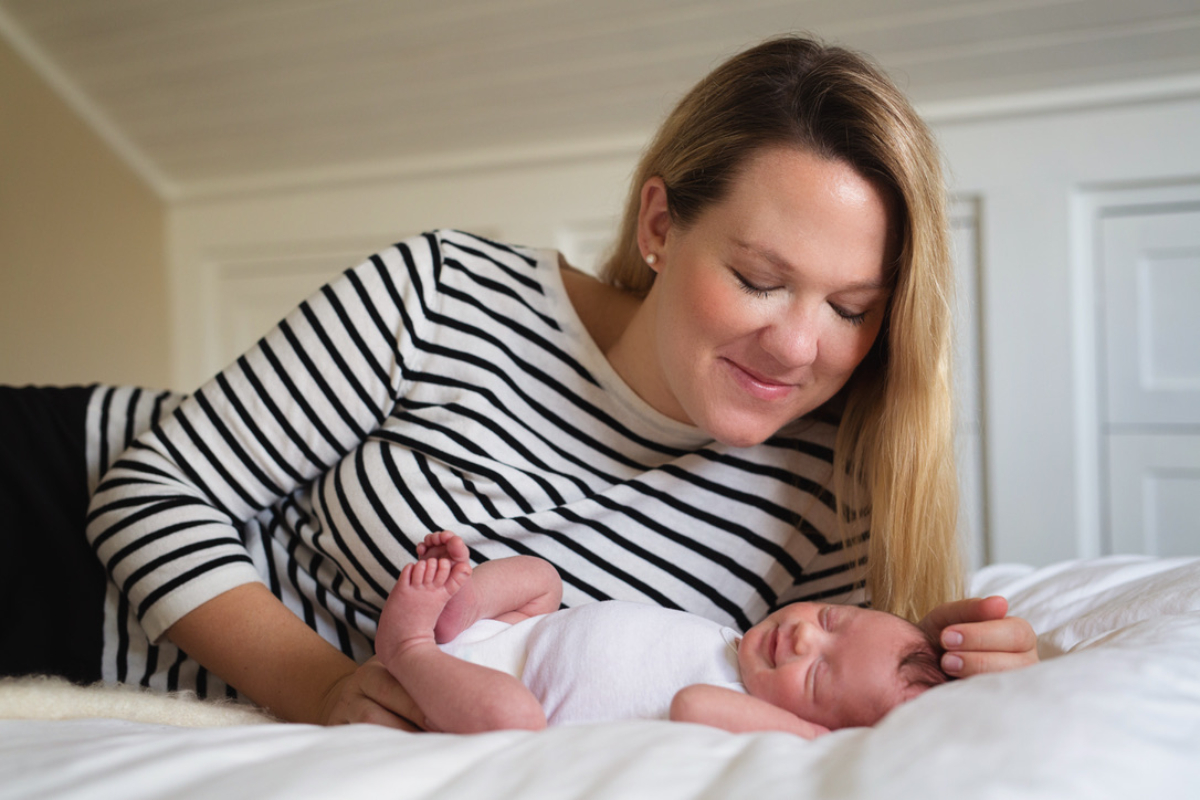 Mother gently strokes her sleeping newborn while relaxing together on a bed during an in-home lifestyle session. This quiet, intimate moment reflects the warmth and connection many families seek before visiting twin cities postpartum cafes for a peaceful outing.