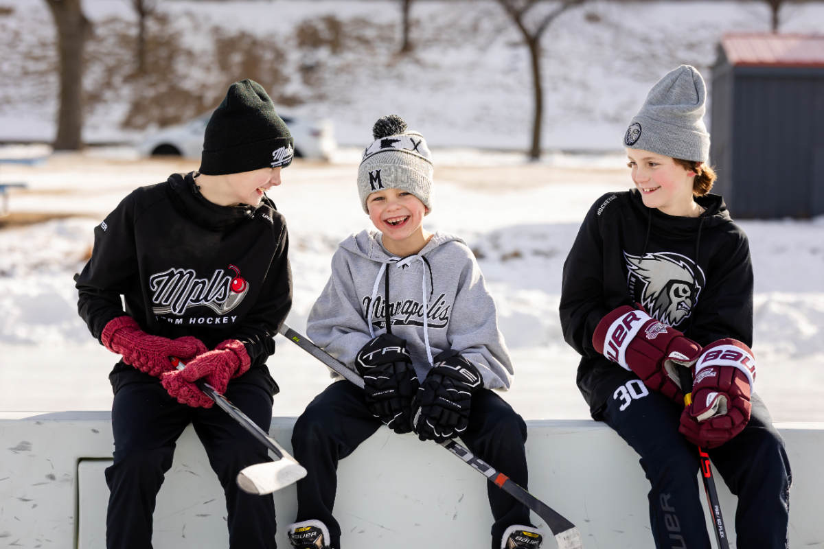 Three brothers in hockey gear sit on the boards at Lake of the Isles rink during a Minneapolis winter family photo session.