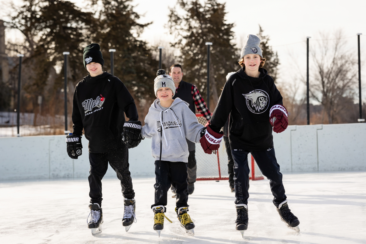 Three brothers hold hands as they skate toward the camera in hockey gear during Minneapolis winter family photos at the Lake of the Isles rink, with their dad skating a few feet behind them.