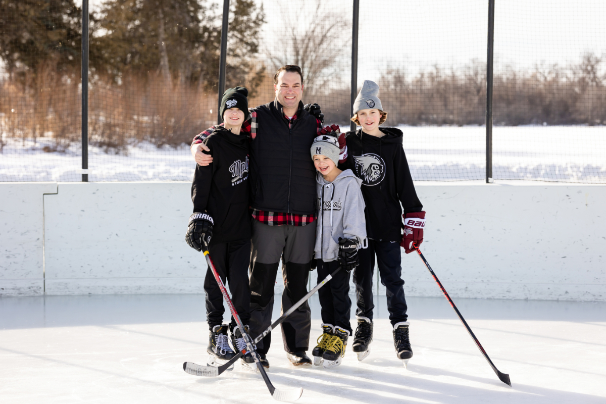 A dad stands arm in arm with his three sons on the ice, all four smiling with hockey sticks in hand, during Minneapolis winter family photos at the Lake of the Isles rink in February.