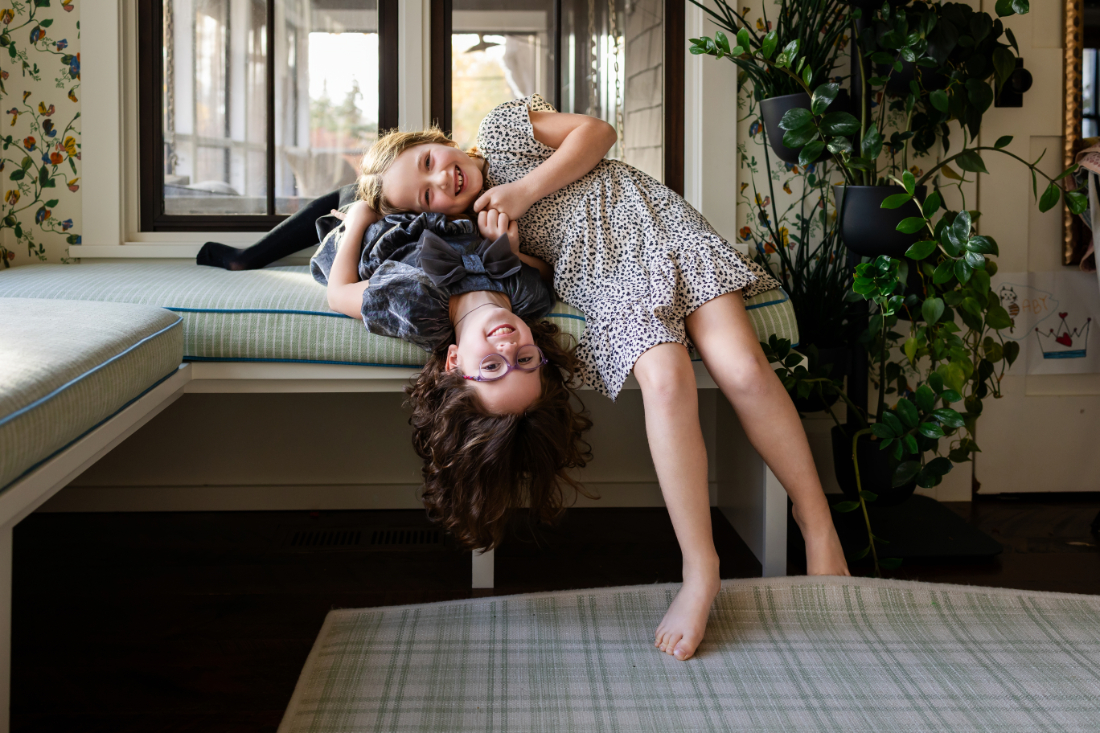 Two sisters laughing and hanging upside down on a window seat, showing storytelling in family photography through playful everyday moments.