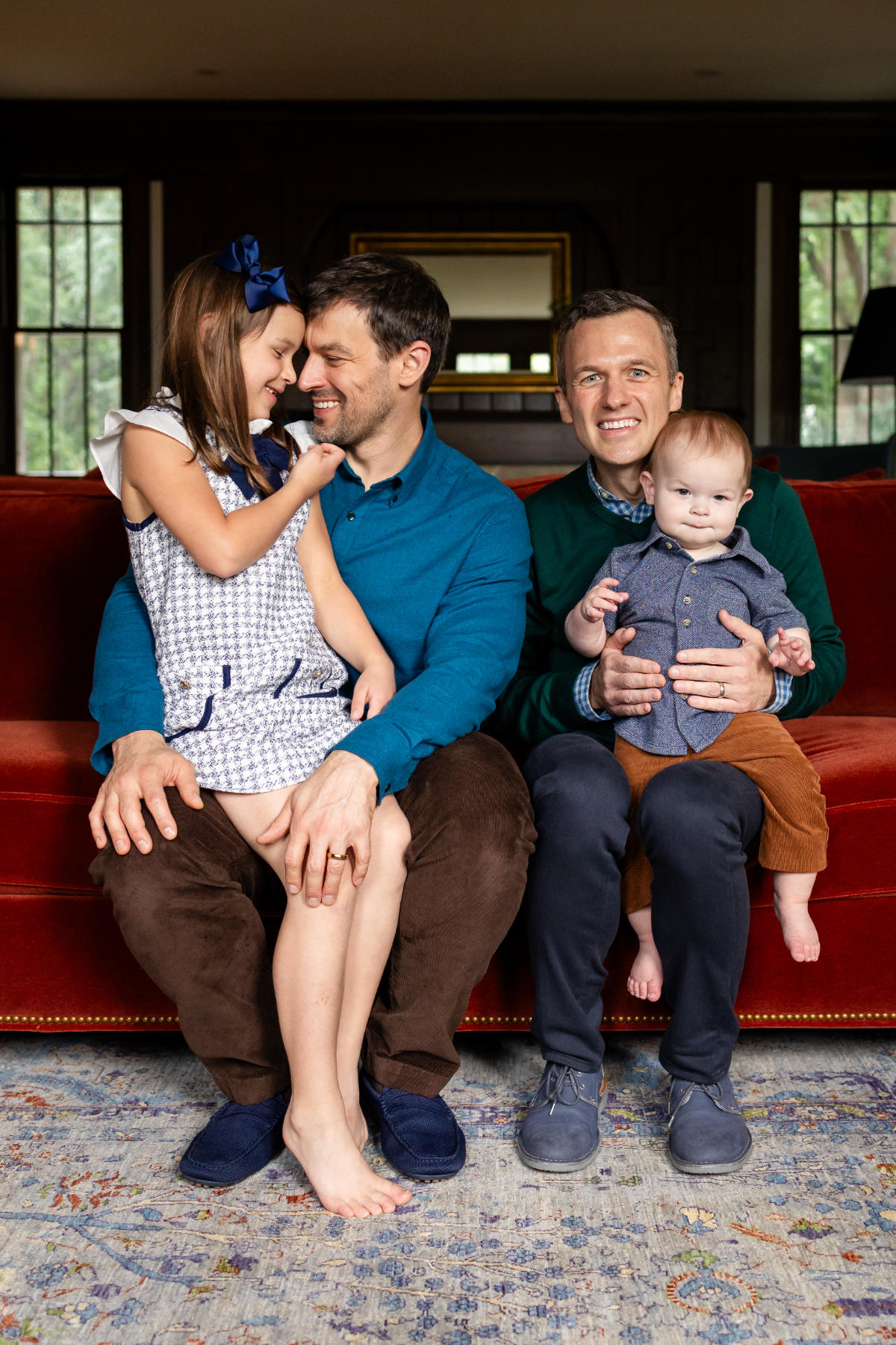 Two dads sit on a red velvet sofa at home with their young daughter and baby son during a Minnesota family photography session. Energy healing supports anyone in a caregiving role, not only moms, at any stage of parenting.