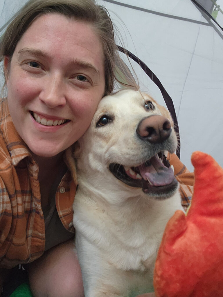 Kayla Kronfeld, a home birth midwife in Minnesota and founder of Meadowlark Midwifery, smiles inside a tent with her yellow lab and a bright orange dog toy. She serves families across the Twin Cities area.