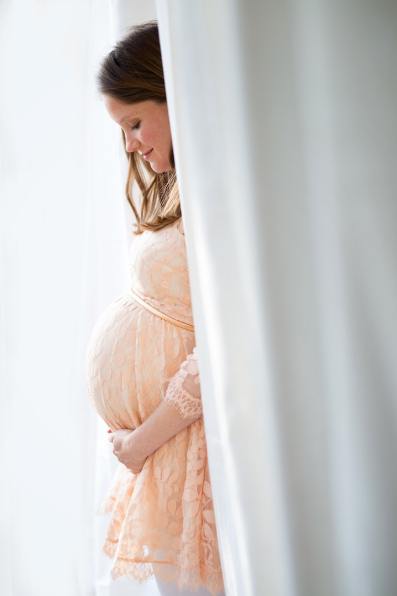 Expecting mom in a soft peach lace maternity dress, standing between white curtains in bright window light with her hands resting under her bump. Soft neutral tones like this photograph beautifully for indoor maternity photos.