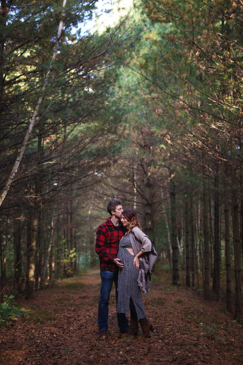Couple on a wooded fall trail in Minnesota. Dad wears a red plaid flannel and kisses mom's forehead while she wears a gray knit dress, tan blanket scarf and brown boots. Coordinated layers, not matching outfits.