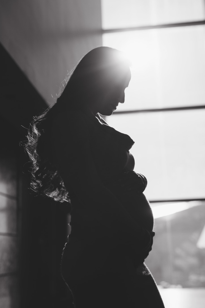 Black and white silhouette of a pregnant woman cradling her bump beside a bright window, backlit by afternoon sun. A dark, simple dress shows how solid colors work for what to wear for maternity photos in Minnesota.