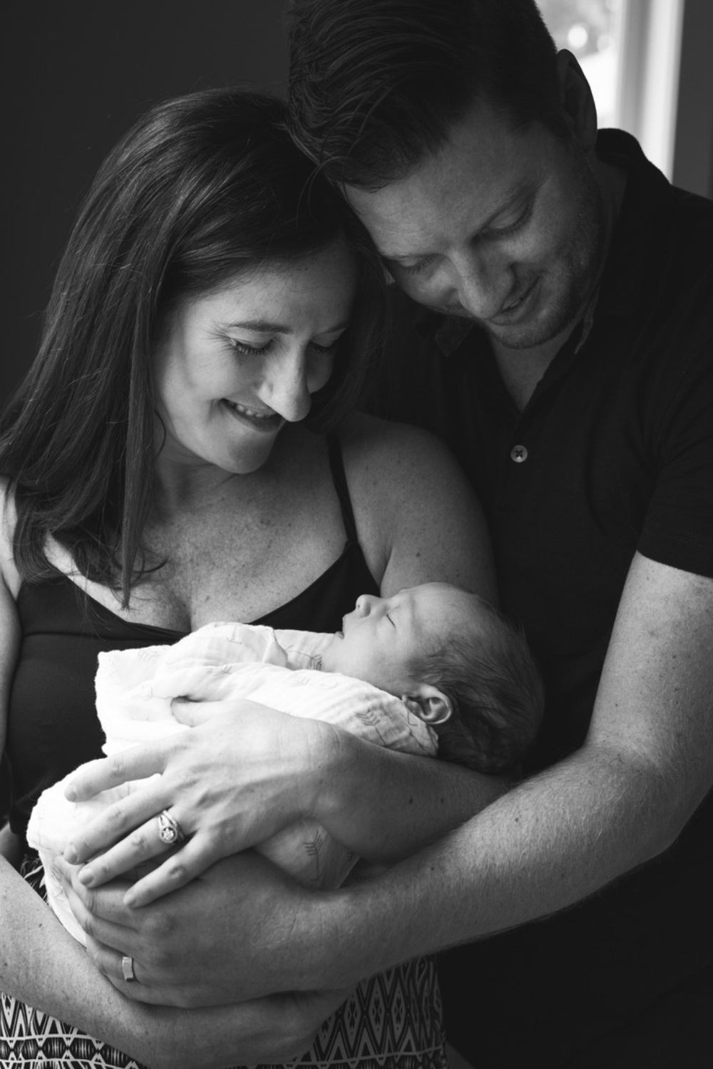 Black-and-white portrait of two parents smiling down at their sleeping newborn, cradled in their arms during an in-home newborn session. Their heads rest close together as they share a quiet moment with their baby.