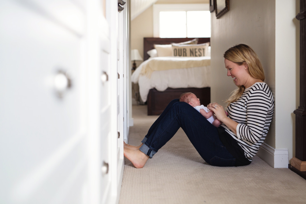 A new mom sits on the carpeted hallway floor, smiling at her newborn resting against her bent knees during an in-home newborn session. Natural light fills the hallway, with the family's bedroom visible in the background.