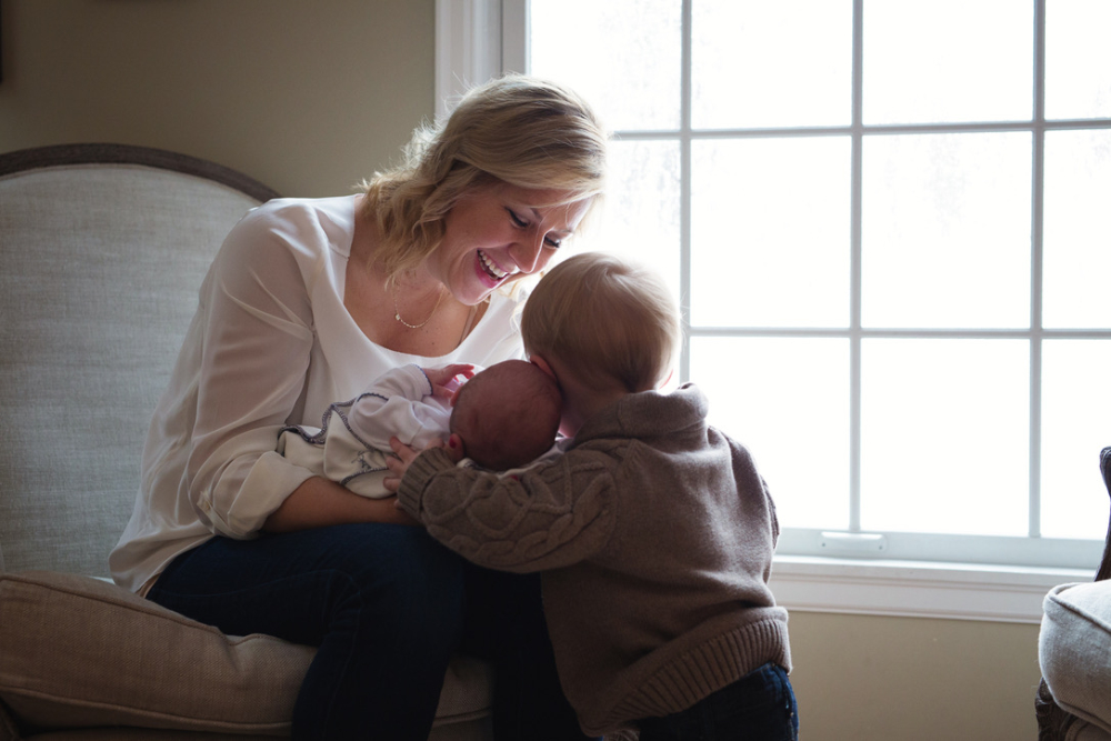 A smiling mom holds her newborn close as an older sibling leans in to kiss the baby's head during an in-home newborn session. The family sits together in a cozy chair beside a large window, surrounded by soft natural light.