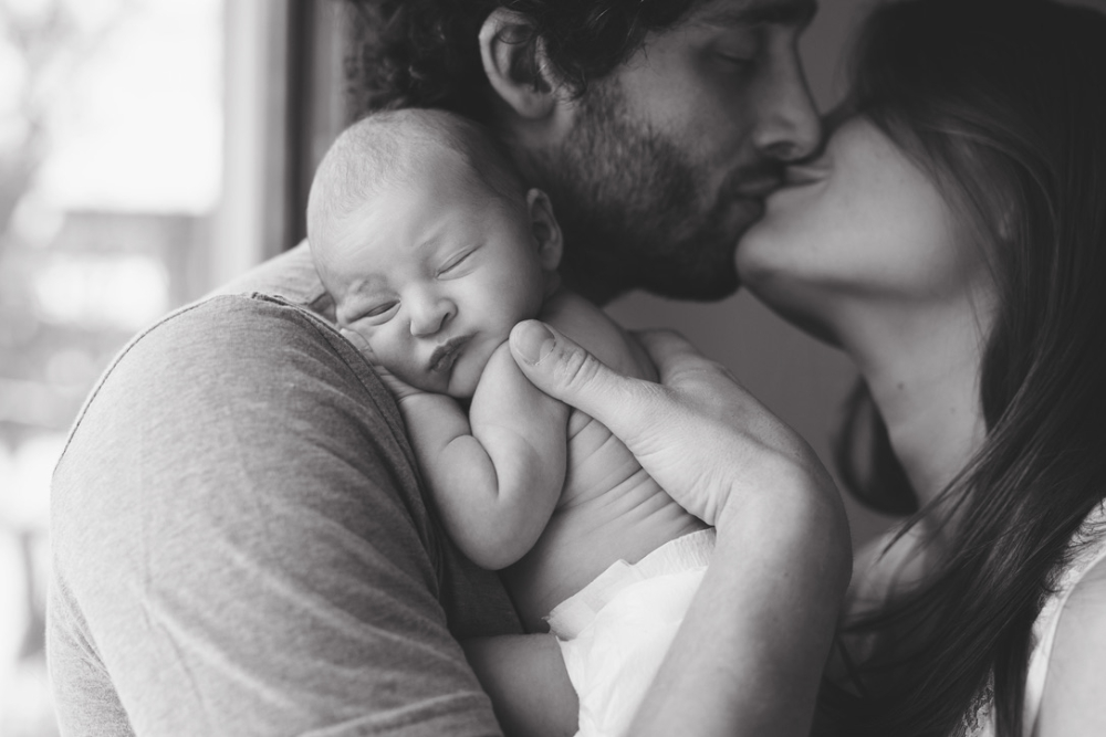 Black-and-white close-up of two parents sharing a kiss while their sleeping newborn rests against dad's shoulder. Mom gently cradles the baby's head, capturing an intimate family moment beside a softly lit window at home.