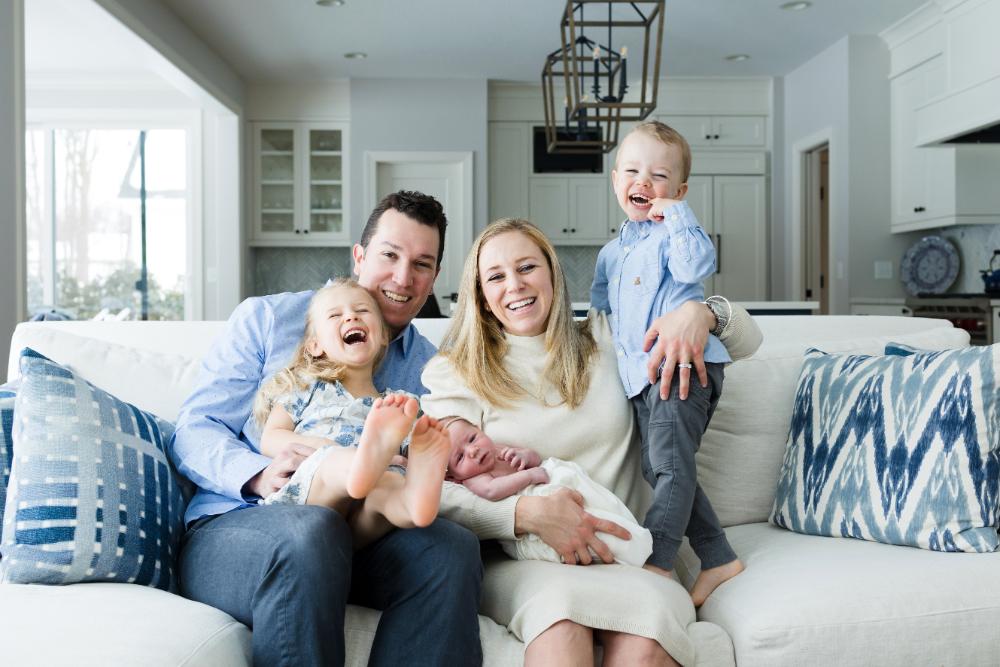 A family of five cuddles together on their living room couch during an in-home newborn session. Mom cradles the newborn while dad holds their laughing daughter, and their older son stands beside them with a big smile.