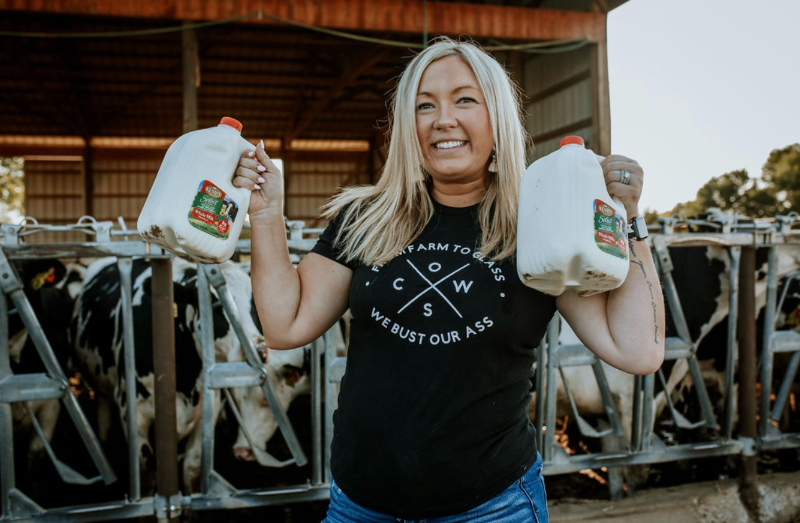 Kennedy Youngren, registered dietitian in Minnesota, smiles holding two gallons of milk in front of dairy cows on her family farm, where she shares postpartum nutrition tips.