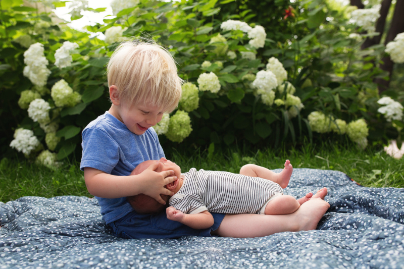 Smiling toddler brother holds his newborn sibling on a blanket by white hydrangeas, a sweet reminder that postpartum nutrition supports the whole family in Minnesota.