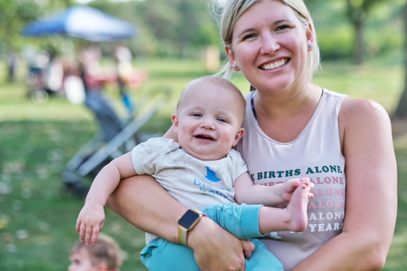 A smiling mom holds her happy baby during an outdoor gathering in a Minnesota park. Moments like these highlight the joy of connecting with other parents and finding motherhood support in Minneapolis through local classes and community groups.