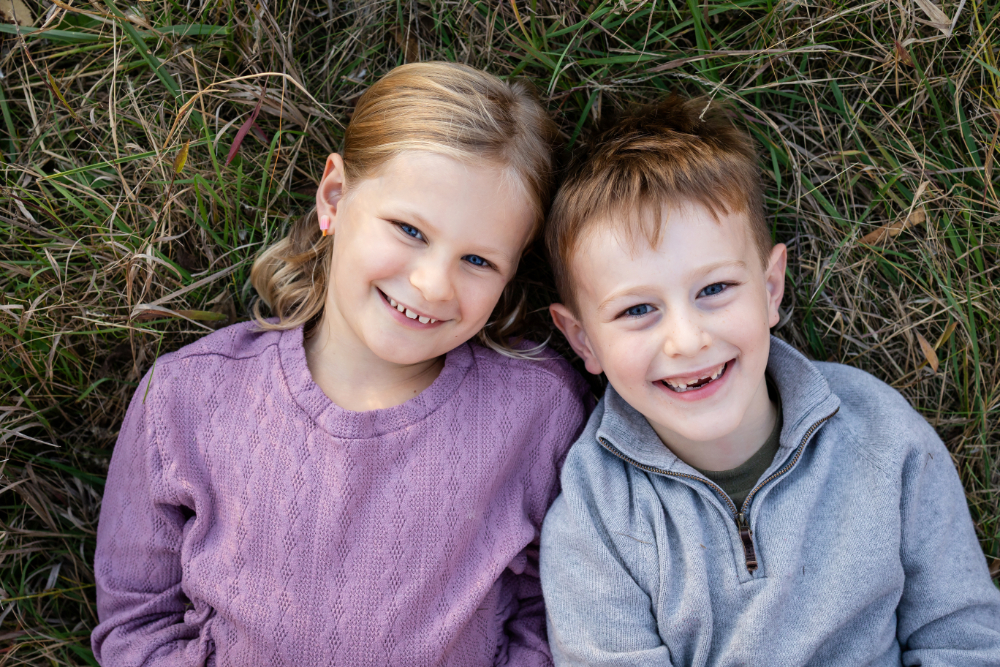 Two smiling children lying together in tall grass, wearing soft purple, gray, and green layers that offer a simple example of what to wear to a photoshoot for relaxed outdoor family pictures.