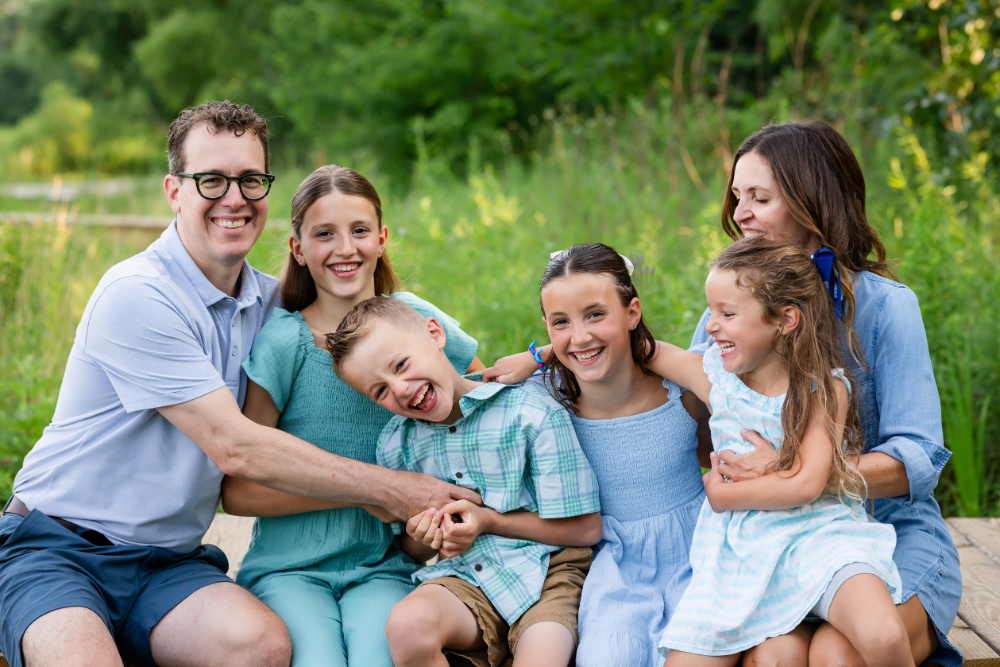 Family of six laughing together outdoors in coordinated shades of blue, green, and white, a great example of what to wear to a photoshoot using complementary colors instead of identical outfits.